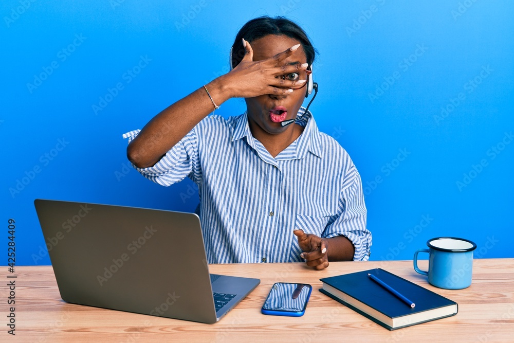 Young african woman wearing call center agent headset peeking in shock covering face and eyes with hand, looking through fingers afraid Young african woman wearing call center agent headset peeking in shock covering face and eyes with hand, looking through fingers afraid
