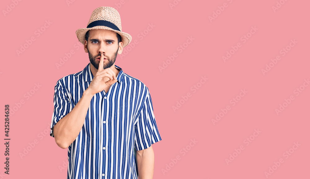 Young handsome man with beard wearing summer hat and striped shirt asking to be quiet with finger on lips. silence and secret concept. Young handsome man with beard wearing summer hat and striped shirt asking to be quiet with finger on lips. silence and secret concept.