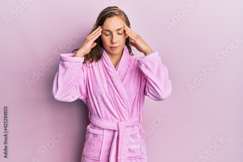 Young blonde woman wearing bathrobe with hand on head for pain in head because stress. suffering migraine.