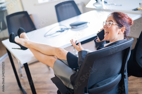 Business woman folded her legs at the office desk and talking on a cell phone. Boredom at work.