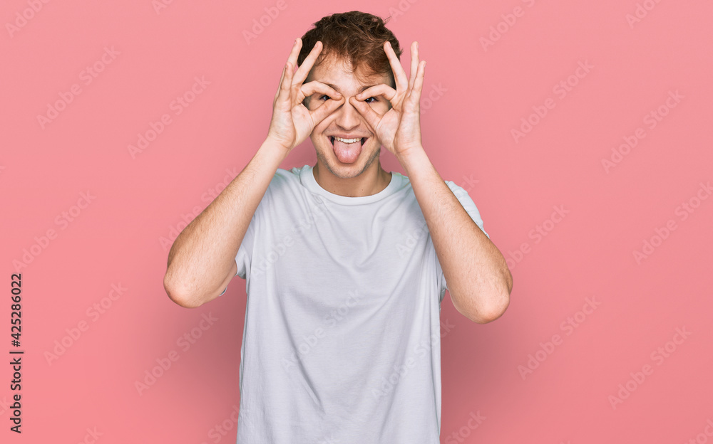 Young caucasian man wearing casual white t shirt doing ok gesture like binoculars sticking tongue out, eyes looking through fingers. crazy expression. Young caucasian man wearing casual white t shirt doing ok gesture like binoculars sticking tongue out, eyes looking through fingers. crazy expression.