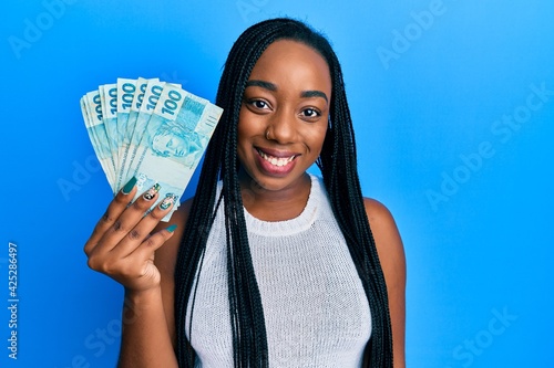 Young african american woman holding 100 brazilian real banknotes looking positive and happy standing and smiling with a confident smile showing teeth
