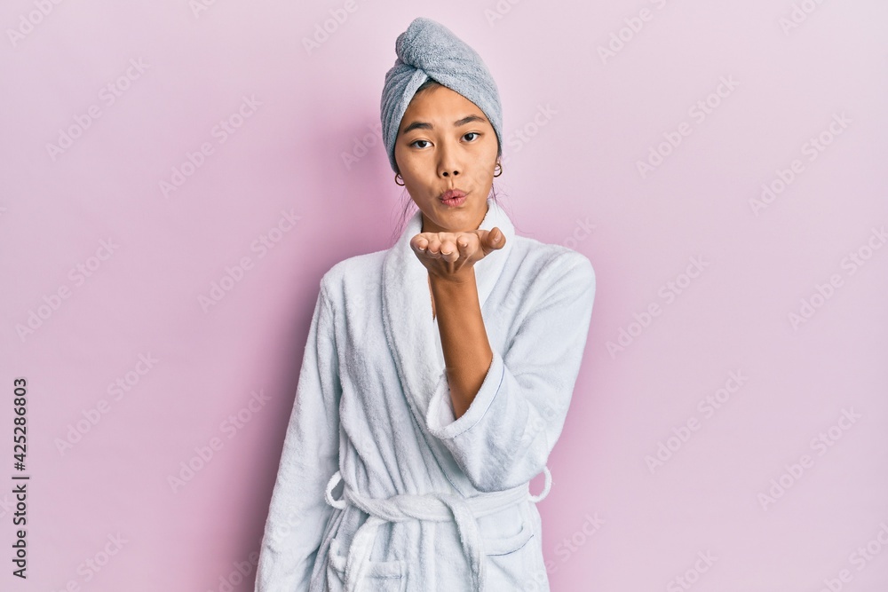 Young chinese woman wearing shower towel cap and bathrobe looking at the camera blowing a kiss with hand on air being lovely and sexy. love expression. Young chinese woman wearing shower towel cap and bathrobe looking at the camera blowing a kiss with hand on air being lovely and sexy. love expression.