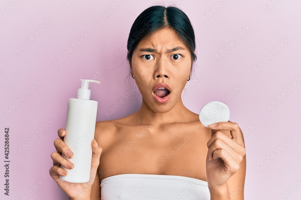 Young chinese woman wearing shower towel using clean lotion in shock face, looking skeptical and sarcastic, surprised with open mouth Young chinese woman wearing shower towel using clean lotion in shock face, looking skeptical and sarcastic, surprised with open mouth