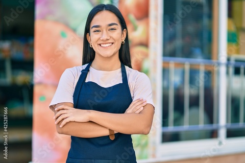Young latin shopkeeper girl with arms crossed smiling happy at the fruit store. photo