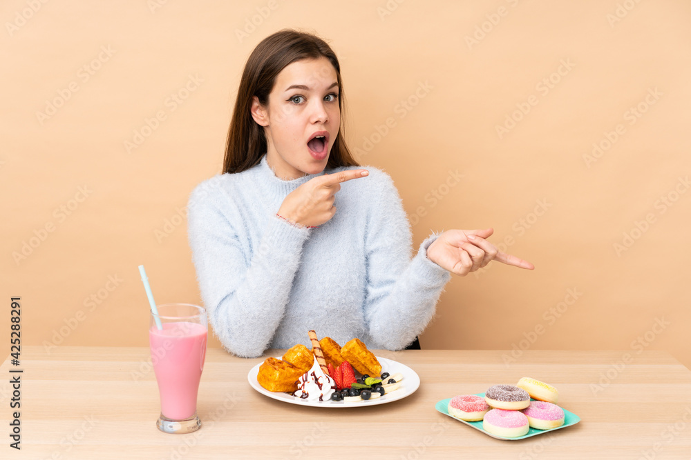 Teenager girl eating waffles isolated on beige background surprised and pointing side Teenager girl eating waffles isolated on beige background surprised and pointing side
