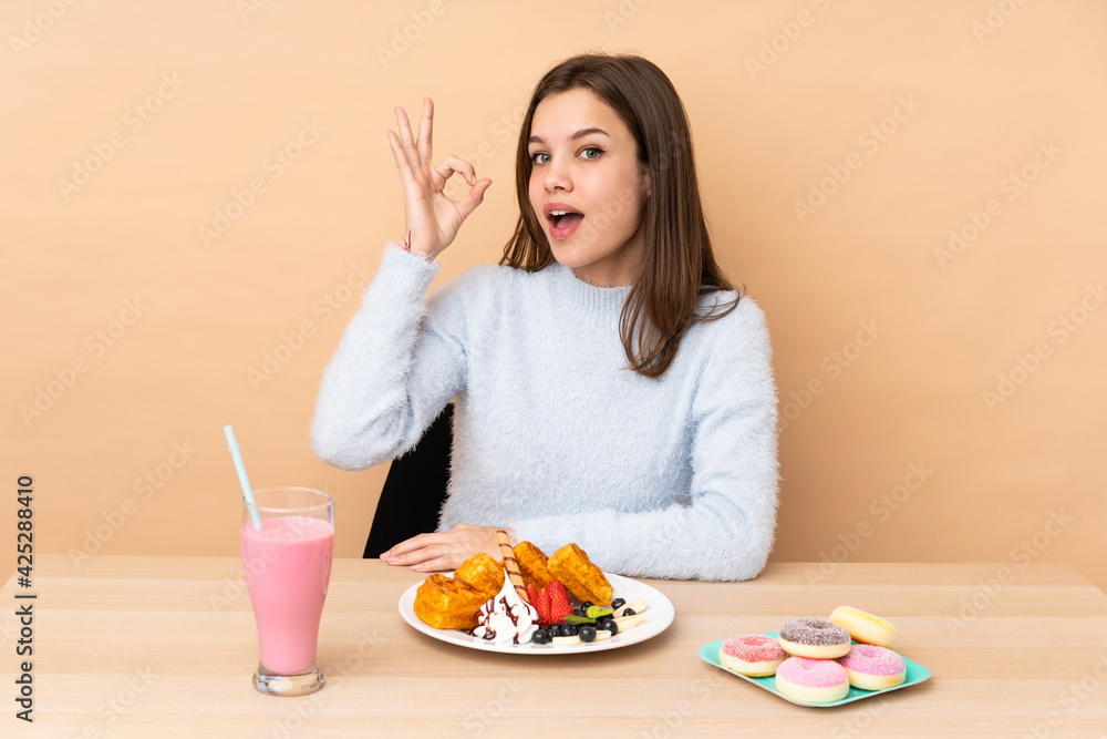 Teenager girl eating waffles isolated on beige background surprised and showing ok sign Teenager girl eating waffles isolated on beige background surprised and showing ok sign