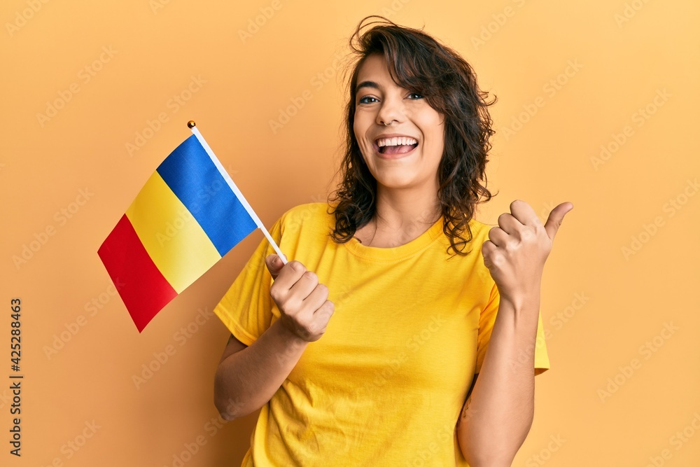 Young hispanic woman holding romania flag pointing thumb up to the side smiling happy with open mouth Young hispanic woman holding romania flag pointing thumb up to the side smiling happy with open mouth