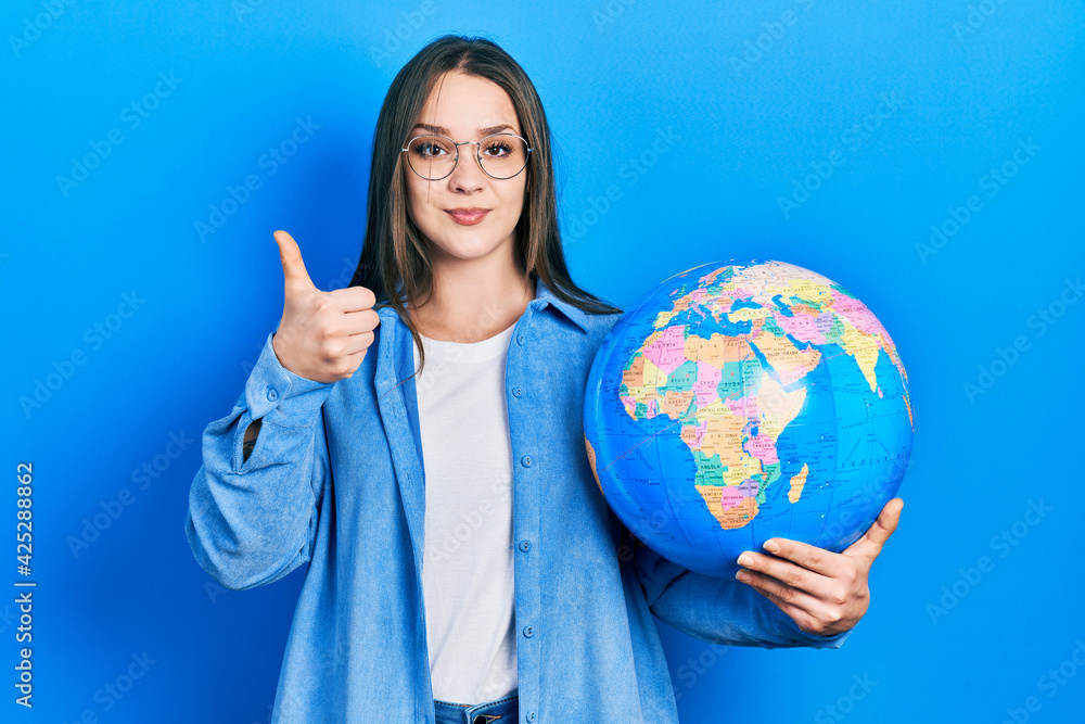 Young hispanic girl holding world ball smiling happy and positive, thumb up doing excellent and approval sign Young hispanic girl holding world ball smiling happy and positive, thumb up doing excellent and approval sign