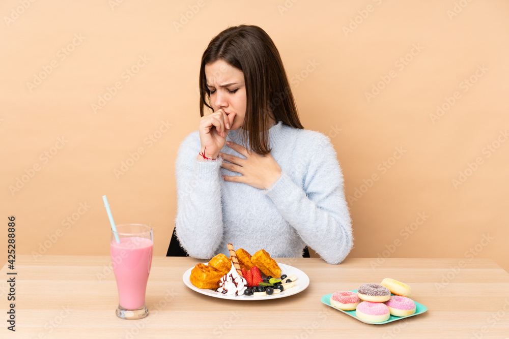 Teenager girl eating waffles isolated on beige background is suffering with cough and feeling bad Teenager girl eating waffles isolated on beige background is suffering with cough and feeling bad
