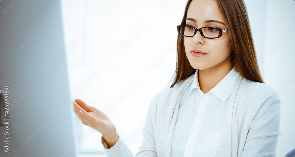 Young woman sitting at the desk with computer in white colored office. Looks like student girl or business lady communicating with casual dressed man Young woman sitting at the desk with computer in white colored office. Looks like student girl or business lady communicating with casual dressed man