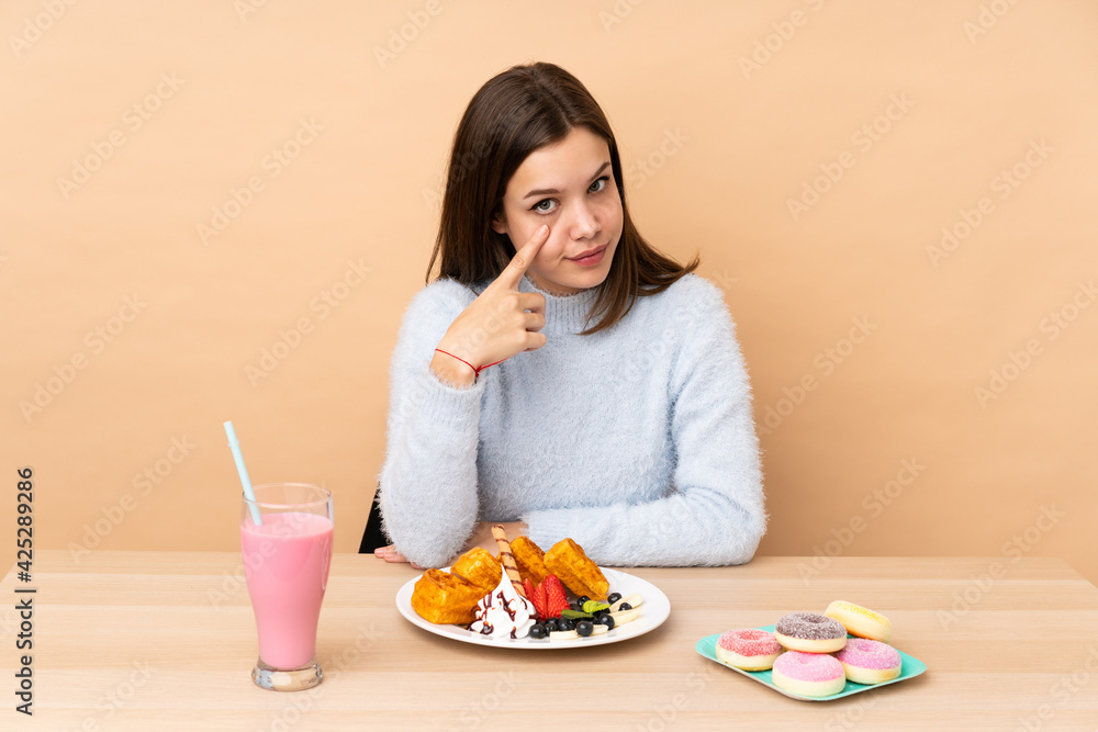 Teenager girl eating waffles isolated on beige background showing something Teenager girl eating waffles isolated on beige background showing something