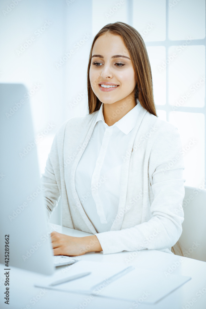 Young cheerful woman sitting at the desk with computer and looking at camera in white colored office. Looks like student girl or business lady. Headshot Young cheerful woman sitting at the desk with computer and looking at camera in white colored office. Looks like student girl or business lady. Headshot