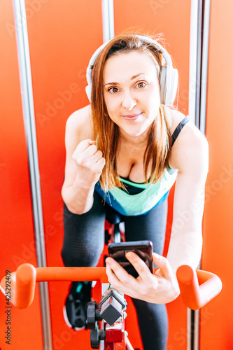 Bild auf Leinwand Young fit athlete woman holding a smart phone while doing biking indor clenching her fist