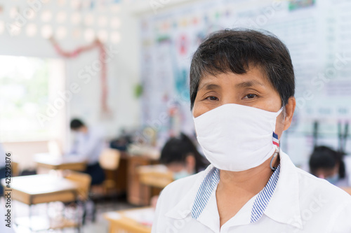 Asian Female teacher wearing white face mask safety during COVID-19 pandemic coronavirus epidemic standing look at camera in front classroom school when all students exam test. Healthcare on Education