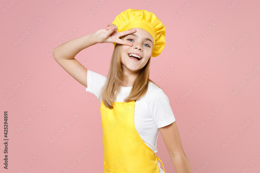 Teen girl chef cook confectioner baker housewife mother's helper in yellow apron white t-shirt cap cover eye with victory v-sign gesture isolated on pink background studio portrait. Food cake concept. Teen girl chef cook confectioner baker housewife mother's helper in yellow apron white t-shirt cap cover eye with victory v-sign gesture isolated on pink background studio portrait. Food cake concept.