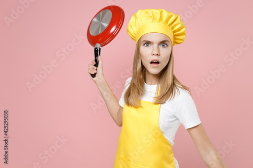Teen angry strict girl chef cook confectioner housewife baker wear yellow apron white t-shirt cap threatening with red frying pan isolated on pastel pink background studio portrait Food cake concept. © ViDi Studio