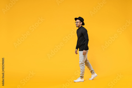 Full length side view young smiling fashionable fun african american man 20s wearing stylish black hat shirt eyeglasses walking going strolling isolated on yellow orange background studio portrait