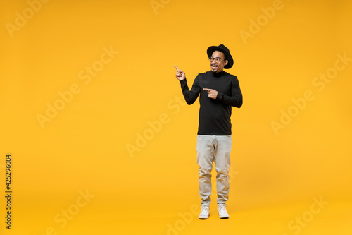 Full length young smiling fashionable fun african american man 20s wear stylish black hat shirt eyeglasses point index finger aside on workspace area isolated on yellow background studio portrait