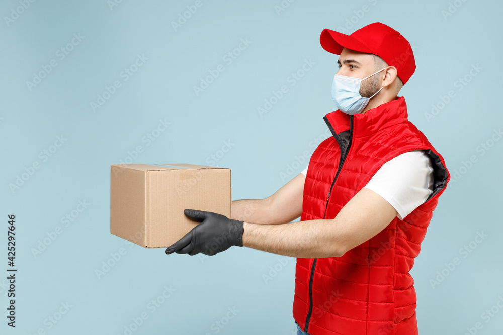 Delivery guy employee man in red cap white t-shirt vest uniform sterile face mask gloves work courier service on lockdown covid19 flu virus hold empty cardboard box isolated on pastel blue background Delivery guy employee man in red cap white t-shirt vest uniform sterile face mask gloves work courier service on lockdown covid19 flu virus hold empty cardboard box isolated on pastel blue background