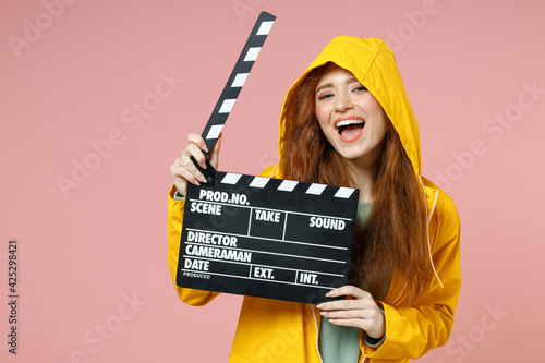 Redhead smiling fun young woman in yellow waterproof hood raincoat outerwear holding classic black film making clapperboard isolated on pastel pink background studio. Lifestyle fall season concept © ViDi Studio