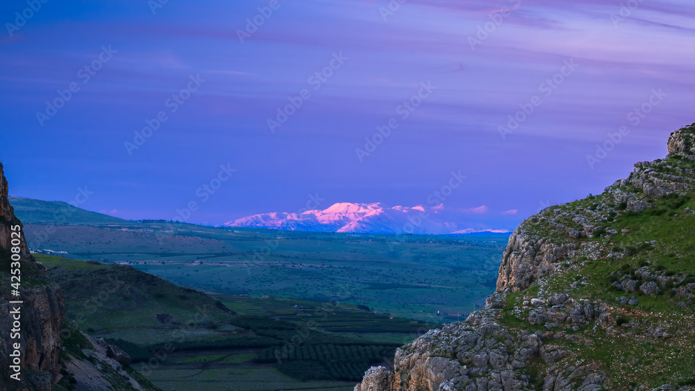 Beautiful view of Galilee from the cliff of Mount Arbel National Park and Nature Reserve, with the snow-capped Mt Hermon lit up by pink sunset light in the distance; Lower Galilee, Israel