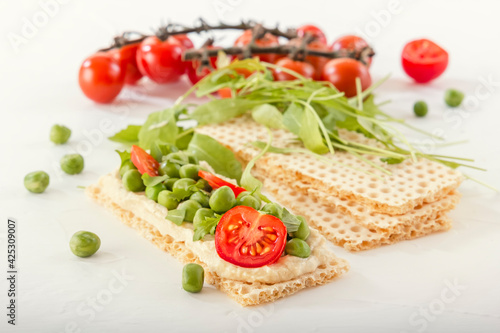 Crispy low calorie wheat crackers with green peas, arugula, microgreens and tomatoes on a white wooden table. Close-up