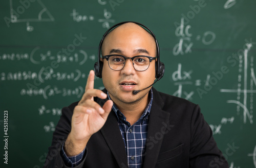 Indian young teacher man looking at camera sitting teaching online video conference live stream. Asian teacher wearing headset teaching class webinar online for students learning.