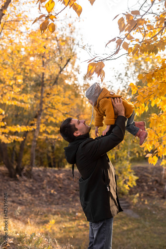 Young handsome father holding his little daughter in an orange coat in his arms in a sunny autumn forest