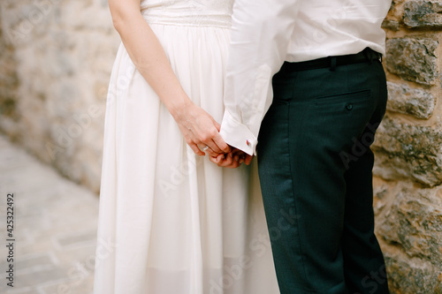 The bride and groom stand near a stone wall and touchingly hold each other's hands, close-up 
