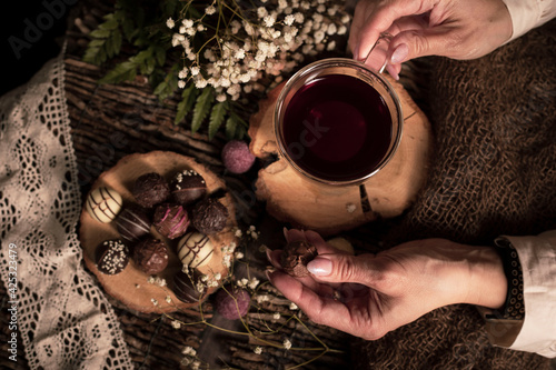 Teezeit in rustikal Still. Schleier Kraut liegt als Deko auf dem Holztisch. Glas Tee steht auf dem Holzhintergrund, bunte Pralinen liegen liegen auf dem Stück Holz. Hände einer Frau, 