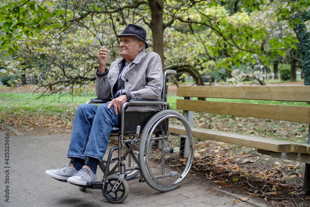 Disabled sad senior man smokes in outdoor while sitting in wheelchair Disabled sad senior man smokes in outdoor while sitting in wheelchair