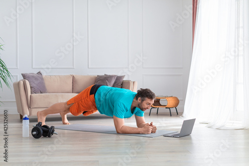 Photo of a middle-aged man during a home workout - he does a plank exercise in front of a laptop monitor.