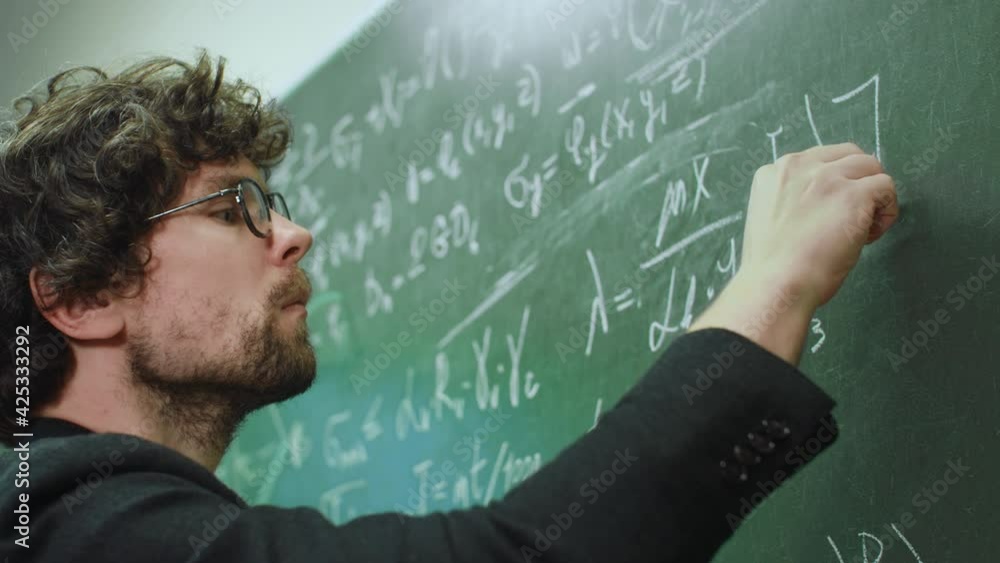 Intelligent stylish teacher wearing eyeglasses writes math formulas with chalk on green chalkboard in university or school classroom. Education and studying concept