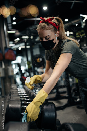 Young female worker disinfecting cleaning and weeping expensive fitness gym equipment with alcohol sprayer and cloth. Coronavirus global world pandemic and health protection safety measures. © Dusko