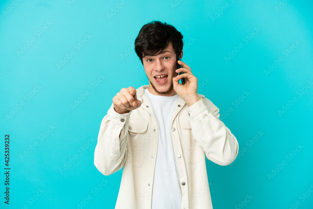 Young Russian man using mobile phone isolated on blue background surprised and pointing front Young Russian man using mobile phone isolated on blue background surprised and pointing front