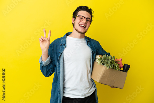 Russian Man making a move while picking up a box full of things isolated on yellow background smiling and showing victory sign