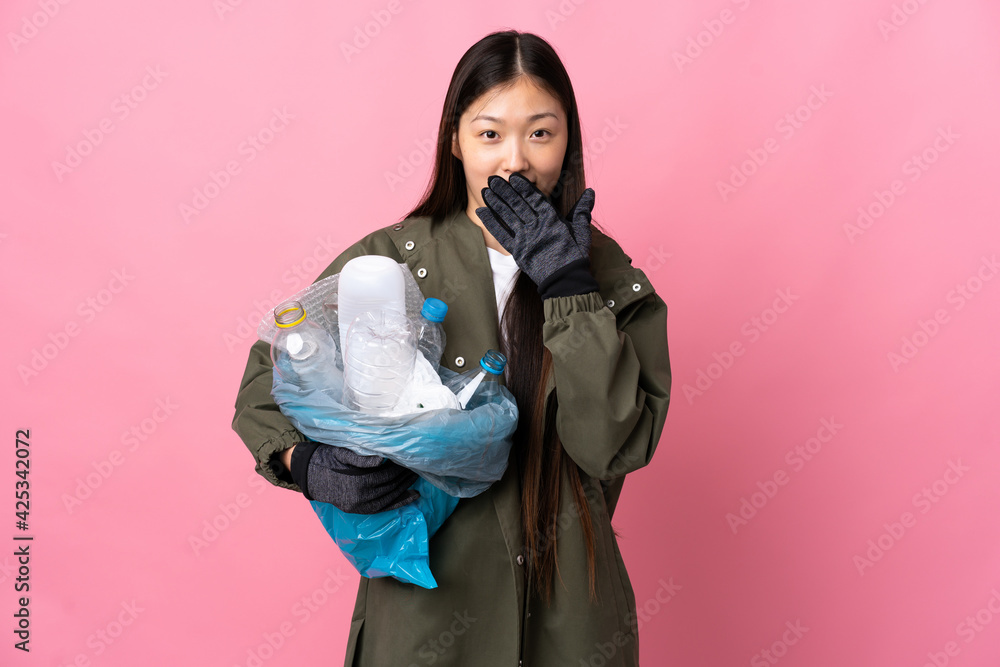 Chinese girl holding a bag full of plastic bottles to recycle over isolated pink background happy and smiling covering mouth with hand Chinese girl holding a bag full of plastic bottles to recycle over isolated pink background happy and smiling covering mouth with hand