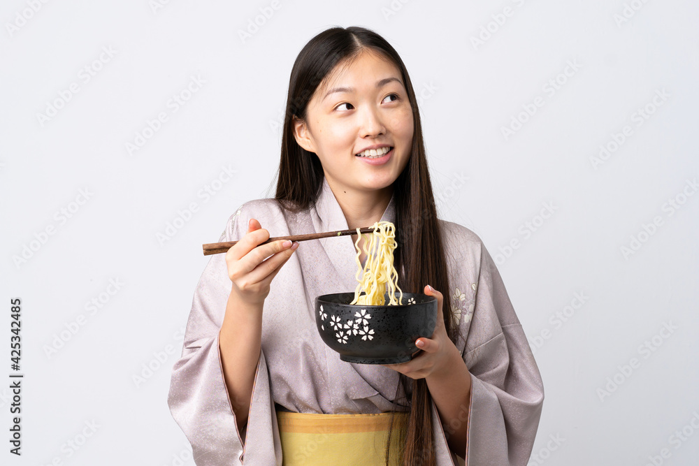 Young Chinese girl wearing kimono over isolated white background holding a bowl of noodles with chopsticks and looking up Young Chinese girl wearing kimono over isolated white background holding a bowl of noodles with chopsticks and looking up