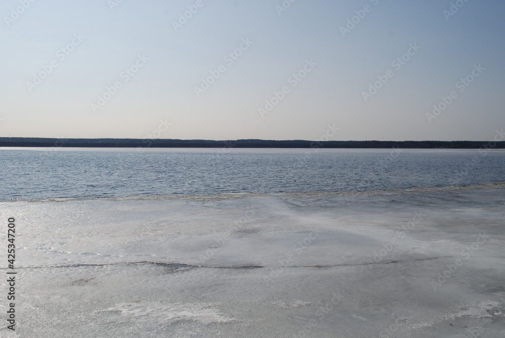 Obraz premium A river is being freed from ice. Spring day light blue sky, river flood with blue water. At the very shore there is a small strip of ice, the middle of the river is without ice. On other side trees