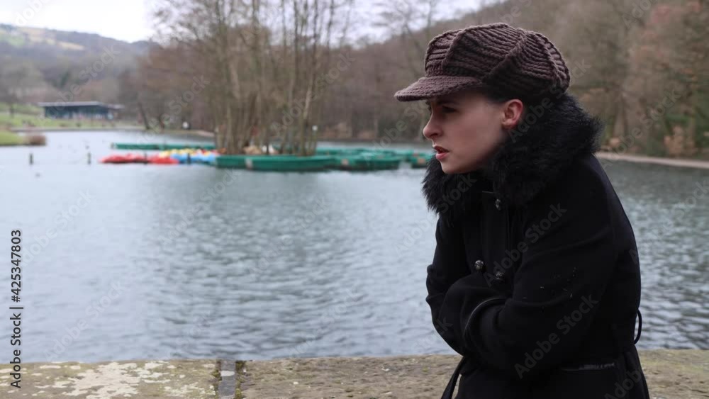 An attractive young woman in a park looking cold and upset while waiting for somebody by a lake in the winter time with a thick black coat and knitted hat