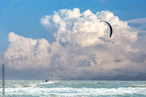 Man kitesurfing with his kiteboard on perfect weather and perfect wind. Big fluffy clouds and mountains in the background.