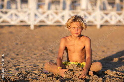 Vászonkép Cute blond teen boy in yellow swim shorts sitting at the sand beach and playing with the sand against white hotel fence
