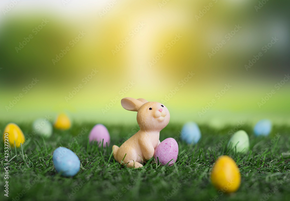 Fototapeta premium Cute little cartoon easter bunny sitting on grass among easter eggs in the garden, easter banner