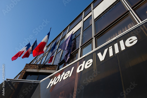 Vue extérieure d'une mairie française avec des drapeaux et le mot 