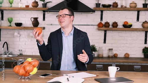 Relaxed male entrepreneur in business suit and glasses tosses an orange from hand to hand while sitting at home in the kitchen during break from work, slow motion. Concept pampering and childishness