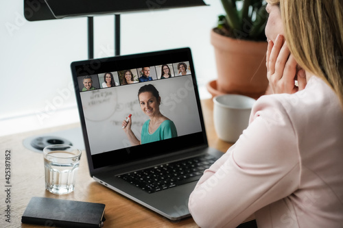 laptop on a work desk showing back view of a woman watching screen of a video conference, a speaker,many attendees, a team meeting or online coaching and worshop event 