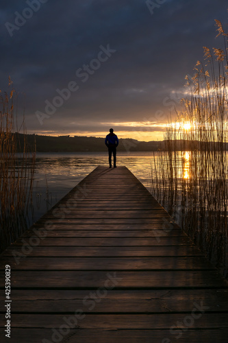 a man stands at the end of an old wooden pier on the shore of a lake at dusk in the golden light. Clouds in the sky and reeds in the water - image