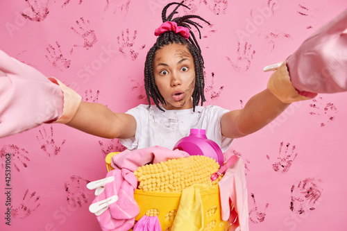 Surprised female model with braids stretches arms takes selfie stares at camera has dirty face and clothes while doing housework busy with household chores poses against pink background near laundry