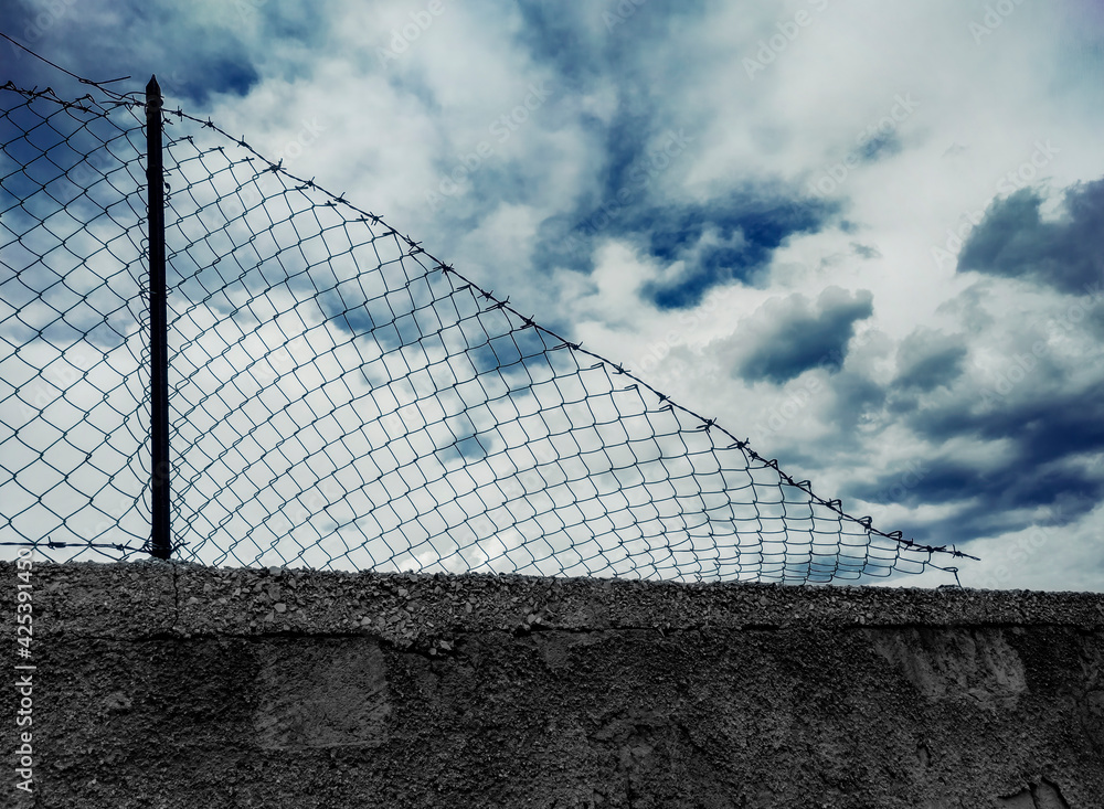 A damaged chainlink fence, with barbed wire on top, installed on a grim gray wall, showing a cloudy but blue sky over it. Symbolic shot: freedom, prison, war, hope, escape.
A damaged chainlink fence, with barbed wire on top, installed on a grim gray wall, showing a cloudy but blue sky over it. Symbolic shot: freedom, prison, war, hope, escape.